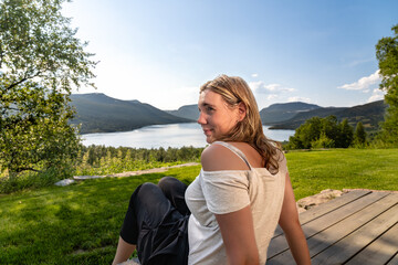 Beautiful woman sits on a wooden deck overlooking the Gjevillvatnet lake, lady looks over her shoulder and past the camera by summer mountain landscape in Oppdal, Tr&oslash;ndelag Norway