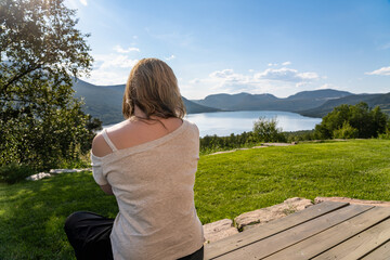 Rear view of a woman sitting on a wooden terrace looking over Gjevillvatnet lake surrounded by the Trollheimen mountain range. The sun shines on her in Oppdal, Tr&oslash;ndelag Norway