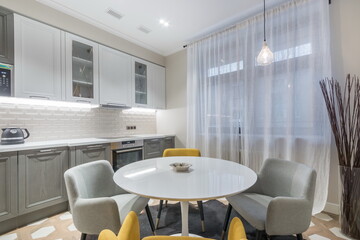 A kitchen is shown with gray cabinets and white countertops. A round table sits in the middle surrounded by soft chairs. Large windows let in natural light