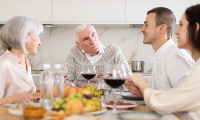 Happy senior parents and young couple talking while drinking wine sitting around kitchen table