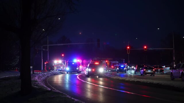 Nighttime scene of multiple emergency vehicles responding at an intersection with flashing lights. Police cars, fire trucks, ambulances, cars stopping at a red light