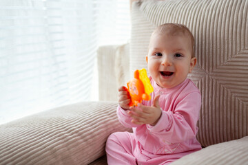 Happy infant baby girl playing on couch at home wearing pink bodysuit. Cute child smiling.