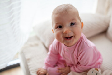 Happy infant baby girl playing on couch at home wearing pink bodysuit. Cute child smiling.
