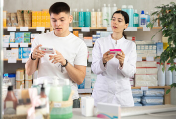 Interested young guy choosing between two boxes of paracetamol antipyretic pills and professional pharmacist woman helping and offering supplements at modern drugstore