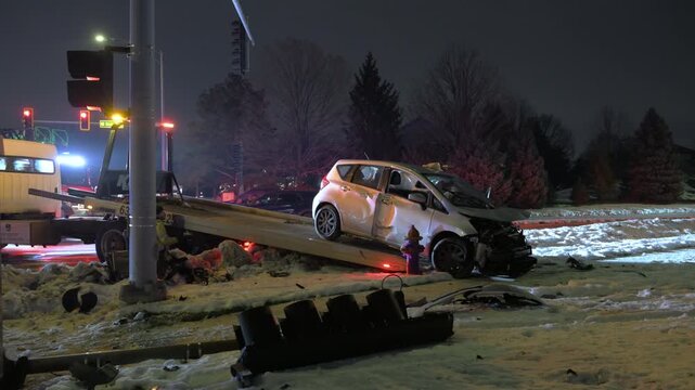 POV damaged compact car being loaded onto a tow truck after a serious accident on a snowy road. Emergency lights illuminate wrecked vehicle, broken traffic pole.