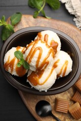 Tasty ice cream with caramel sauce and mint in bowl on black table, flat lay
