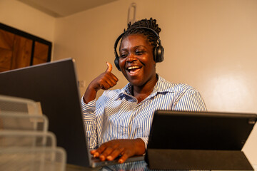 Young African woman with headphones gives a thumbs-up while working on her laptop, involved in...