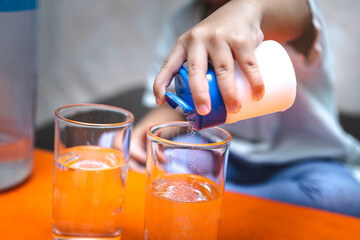 Close-up of a child's hand pouring salt into a glass water from a blue and white container. Concept of nutrition, science experiments, or healthy rehydration for kids at home.