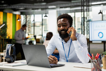 Male manager speaking with business associate on mobile device and using laptop for work. Smiling sales executive multitasking in office, combining phone conversation with digital project planning.
