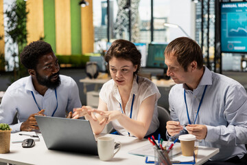 African American and Caucasian colleagues sitting together in modern office, reviewing financial charts on laptop. Male and female employees showcasing teamwork, productivity and corporate strategy.