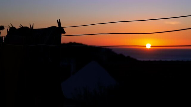 Scenic ocean sunset with silhouetted hut and clothesline. Glowing sun slowly sinking below the ocean horizon, creating a vibrant orange and purple sky with a silhouetted rural hut and clothesline