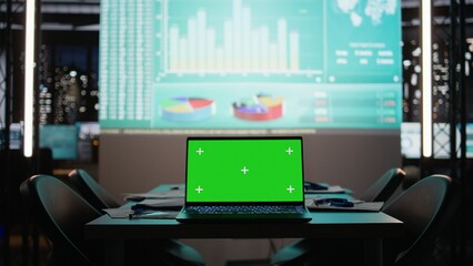 Empty boardroom shows a laptop with copy space screen late at night, illuminated by projected business data and charts. Dark office corporate environment with planning and forecasting.