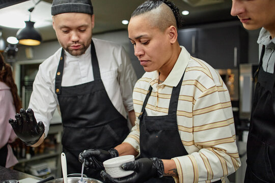 Young adult Caucasian man wearing chef uniform instructing young adult biracial woman preparing food in professional kitchen while another young adult Caucasian man observing