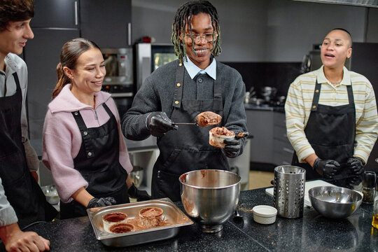Group of young adults preparing chocolate dessert together in kitchen, Black man scooping batter into baking dish while others watching and smiling