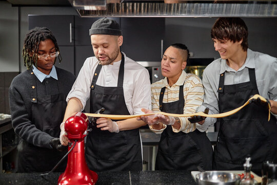 Multiethnic group of young adult men and women making fresh pasta together in kitchen, using stand mixer and working as team, focusing on collaborative cooking process