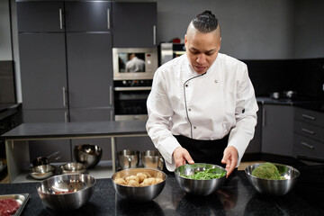 Biracial chef preparing fresh salad ingredients in professional kitchen, arranging vegetables in stainless steel bowls on countertop, focused on culinary task