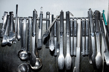 Assorted stainless steel kitchen utensils including ladles, tongs, and knives hanging on metal rack against wall, professional culinary tools organized for easy access in commercial kitchen