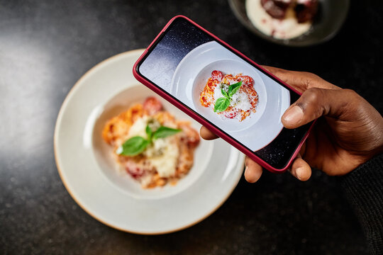 Black man photographing plate of Italian pasta with smartphone, capturing close up of food garnished with basil and grated cheese, hand holding phone above dish on dark table