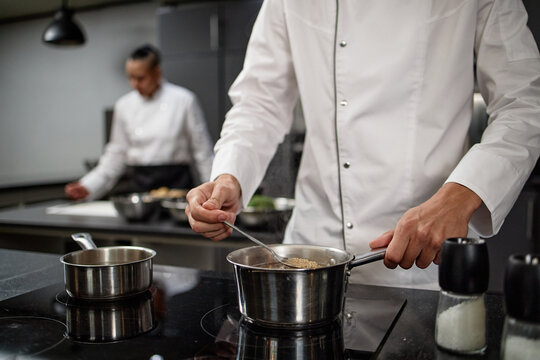 Young adult man stirring food in saucepan on stove in professional kitchen while female chef working in background preparing ingredients