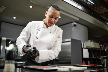Young biracial female chef seasoning raw meat with pepper grinder in professional kitchen, wearing chef uniform and black gloves, preparing ingredients for cooking on stainless steel counter