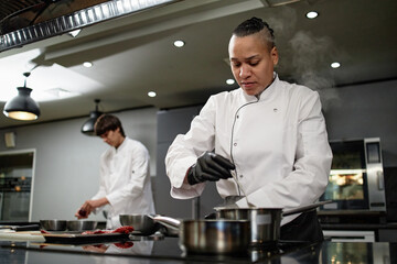 Biracial chef preparing food in professional kitchen, stirring pot on stove while young Caucasian man working in background, both focused on culinary tasks, wearing chef uniforms