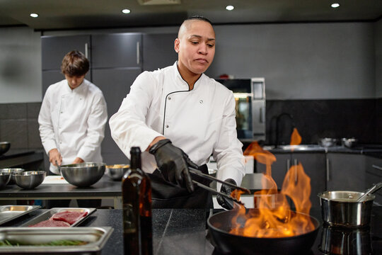 Caucasian young adult man and biracial woman working in professional kitchen, woman grilling food over open flame while man preparing ingredients in background