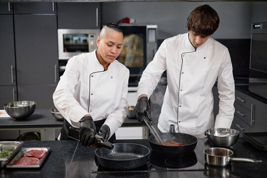 Biracial woman and man cooking together in professional kitchen, both wearing chef uniforms and preparing meat in frying pans on stovetop