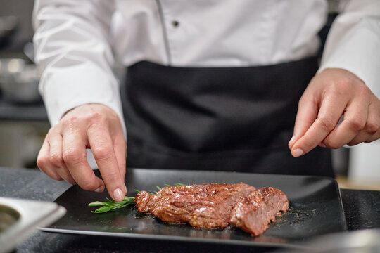 Chef preparing steak on black plate, garnishing cooked meat with fresh herbs in professional kitchen, hands visible arranging food for presentation
