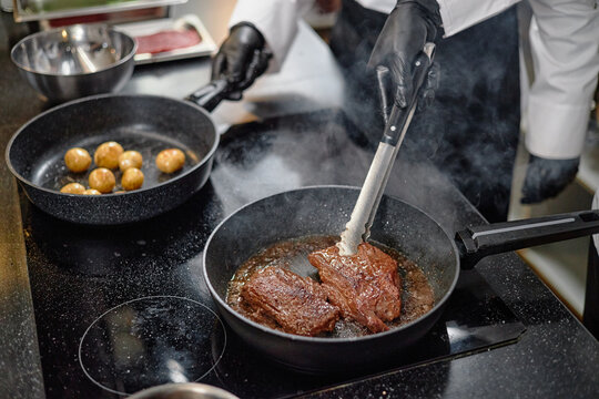 Man wearing chef uniform and black gloves searing steak in frying pan while turning meat with tongs, cooking small potatoes in separate pan on stovetop