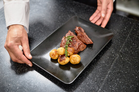 Hands serving grilled steak with roasted potatoes and fresh rosemary on rectangular black plate, presenting dish on dark countertop in restaurant kitchen setting