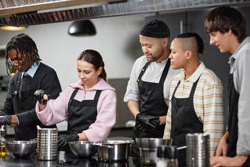 Group of young adult multiethnic men and woman cooking together in professional kitchen, chef supervising as participants preparing ingredients and learning culinary techniques