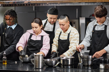 Diverse group of young adult men and women preparing food together in kitchen, wearing aprons and gloves, mixing ingredients in metal bowls, focused on cooking task