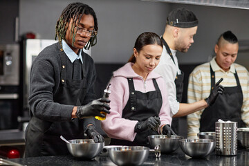 Group of young adult multiethnic men and women preparing food together in kitchen, wearing aprons and gloves, mixing ingredients in metal bowls, collaborating on cooking task