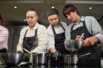 Three diverse young adults wearing aprons cooking together in kitchen, stirring ingredients in metal bowls and saucepans, collaborating on meal preparation
