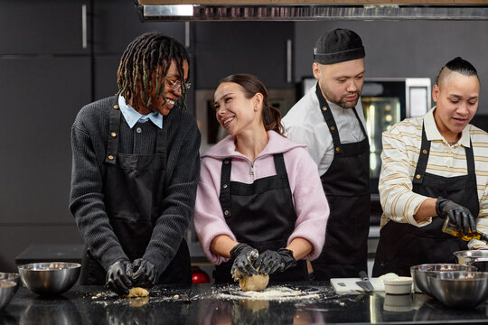 Multiethnic group of young adults preparing dough together in kitchen, smiling and interacting while wearing aprons and gloves, standing at counter with baking ingredients visible