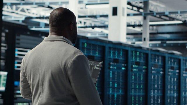 Engineer walking in server farm, automating system diagnostics using machine learning predictions. IT specialist working in data center using notebook to configure gear for AI tasks, camera B