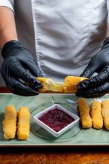 Chef Hands Wearing Black Gloves Breaking Fried Mozzarella Cheese Stick to Show Melted Texture Over Green Plate and Red Sauce
