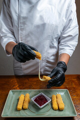Chef Hands Wearing Black Gloves Breaking Fried Mozzarella Cheese Stick to Show Melted Texture Over Green Plate and Red Sauce