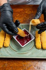 Chef Hands Wearing Black Gloves Breaking Fried Mozzarella Cheese Stick to Show Melted Texture Over Green Plate and Red Sauce