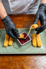 Chef Hands Wearing Black Gloves Breaking Fried Mozzarella Cheese Stick to Show Melted Texture Over Green Plate and Red Sauce