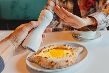 Person's hand making a stop gesture, refusing a pepper mill held by another hand, above a plate of Khachapuri