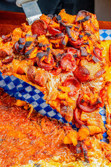 Chef in black uniform and gloves lifting a slice of pizza topped with crispy pork rinds, ripe plantain, and shredded beef, using a spatula on top of various pizza varieties on checkered paper.