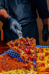 Chef in Black Uniform and Gloves Lifting a Loaded Pepperoni Pizza Slice with Spatula over Diverse Pizza Varieties on Checkered Paper