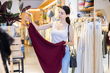 Fototapeta premium Woman is standing in a store near a clothes rail and choosing a stylish summer dress.