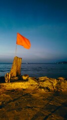 At dusk, a red flag hangs near the rocky shore, prohibiting swimming in the sea.