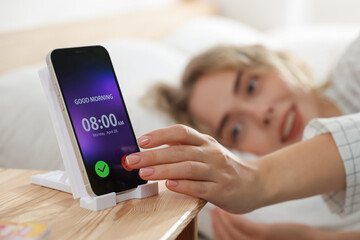 Woman turning off alarm clock on smartphone in bed indoors, selective focus