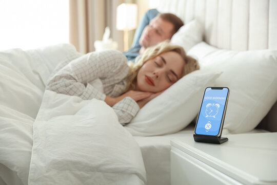 Couple sleeping in bed and smartphone with alarm clock app indoors, selective focus