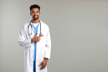 Smiling doctor in medical uniform with stethoscope pointing at something on grey background. Space for text