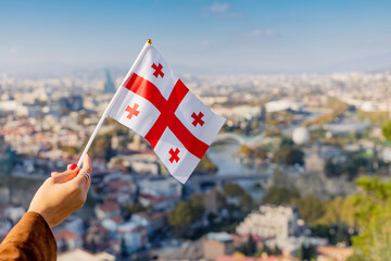 Woman's hand holding a small Georgian flag, symbolizing patriotism and travel, overlooking the panoramic Tbilisi cityscape © EdNurg