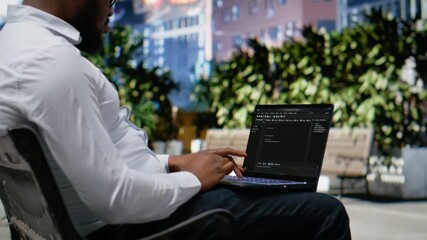 Confident adult reviews binary code script for his programming remote work, sitting on a bench in the city center and working on laptop. Scrolling and typing near lively downtown scenery. Camera B. - Powered by Adobe